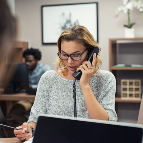 Woman with glasses speaking on the phone at a desk while reviewing documents